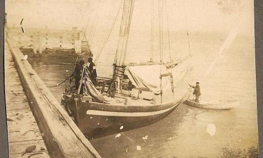 Two men on a wooden sailing ship moored at a wharf. Another man stands in a boat next to the ship.