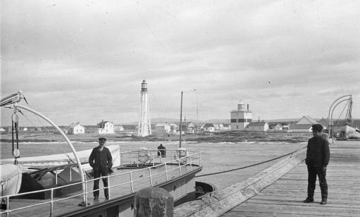 A man stands in a moored boat while another stands on the wharf. A few homes and two lighthouses can be seen in the distance.

