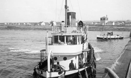 A steamboat approaches a wharf, with another boat behind it. Houses on the shore can be seen in the background. 