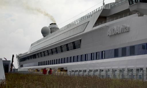 Thick grey smoke rises from one of the funnels of a large white cruise ship moored at a wharf.