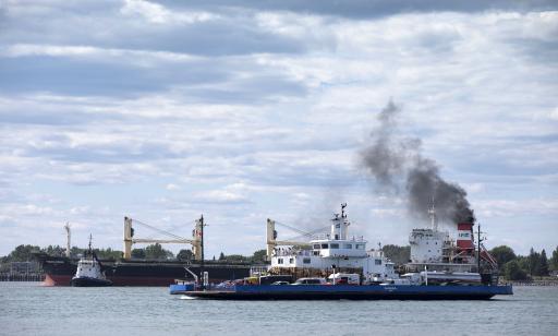 A ferry loaded with cars and trucks passes in front of a cargo ship. Thick black smoke pours from the cargo ship's stack.