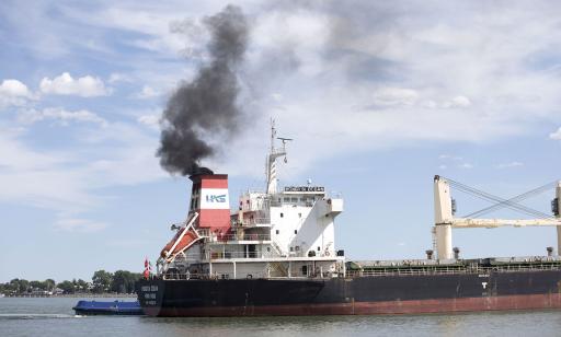Very thick black smoke rises from a cargo ship's stack. A park with its many trees can be seen in the background. 