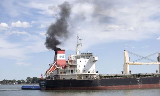 Une abondante fumée noire très opaque sort de la cheminée d’un cargo et, au loin, on voit beaucoup d’arbres dans un parc.