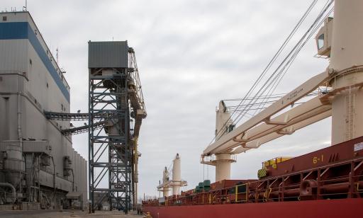 Men stand under a metal tower near some silos on a wharf partially covered in grain. A cargo ship is moored alongside. 