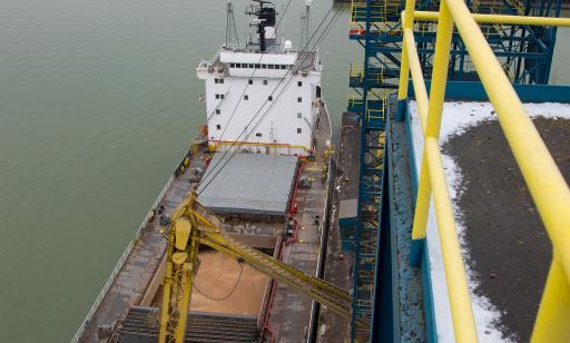 A yellow metal extraction arm entering the hold of a bulk carrier moored at a wharf. The hold is full of yellow grain.