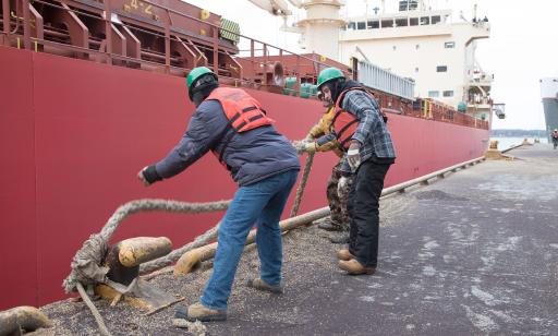 Three workers wearing life jackets and hardhats stand on a wharf, loosening the mooring lines of a bulk carrier.