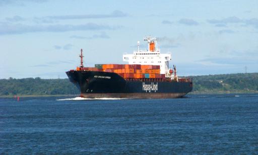 A large ship carrying many metal boxes kicks up a large wake as it passes near the shore.