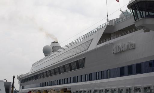 A man and a woman stand on a wharf near a large white ship. Thick grey smoke rises from the ship's stack.