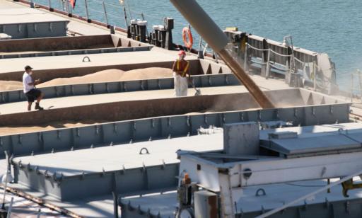 Two men stand on the deck of a large ship, watching as grain pours out of a large tube into a hold. 