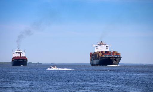 Two large ships loaded with metal boxes sail in the same direction, while a small motorboat crosses their path. 