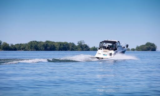 A man in a small yacht speeds past a tree-covered island, leaving a large wake.