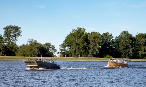 A small, flat motorboat and a dismasted motor-powered sailboat kick up waves as they sail past an island.